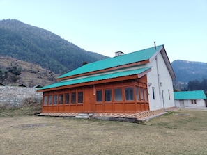 A two-story building with a green roof, wooden exterior, and a stone wall in the background. - Forest Rest House Jibhi (Jibhi)