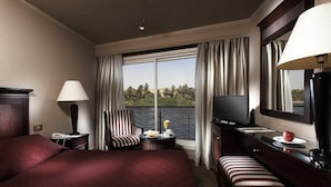 A hotel room with a large bed, a desk with a TV, a chair, a small table with a lamp, and a view of the outdoors. - JAZ Crown Jubilee Nile Cruise - Thursday VIA Luxor 7 & 4nts - Monday VIA Aswan 3nts (Luxor)