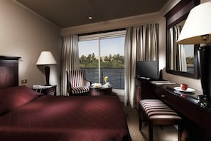 A hotel room with a large bed, a desk with a TV, a chair, a small table with a lamp, and a view of the outdoors. - JAZ Crown Jubilee Nile Cruise - Thursday VIA Luxor 7 & 4nts - Monday VIA Aswan 3nts (Luxor)