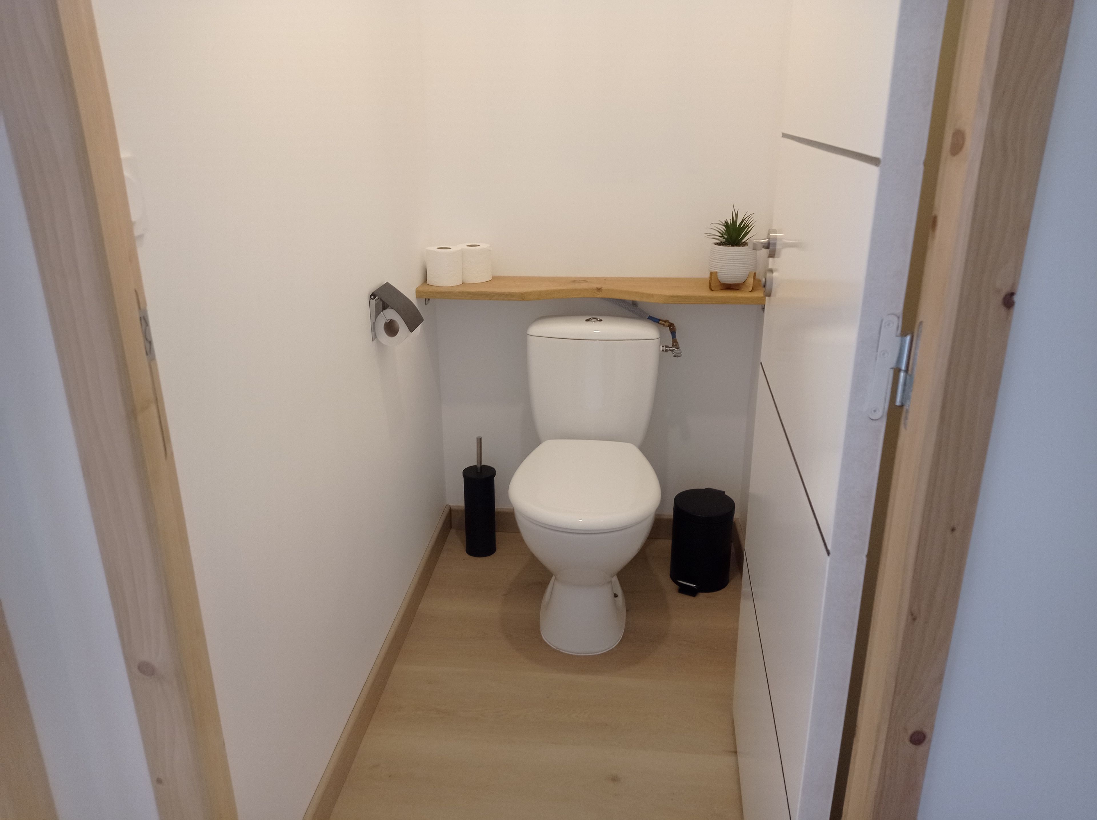 A small, narrow bathroom with a toilet, a toilet paper holder, a trash bin, and a small shelf with a plant.