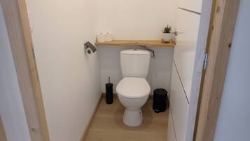 A small, narrow bathroom with a toilet, a toilet paper holder, a trash bin, and a small shelf with a plant.