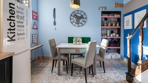 Een keuken met een eettafel en stoelen, een hanglamp, een klok en een plank met keukengerei.