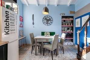 Une cuisine avec une table à manger et des chaises, un luminaire suspendu, une horloge et une étagère contenant des ustensiles de cuisine.