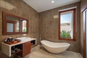 A modern bathroom with a freestanding bathtub, a vanity with a mirror, and a window with a view of greenery. - Monaco Blu Luxury Villas and Spa (Keroboken Kelod)