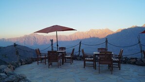 A terrace with stone flooring, wooden chairs, and tables, surrounded by a rope barrier, overlooking a mountain range. - Sama Wakan (Wakan)