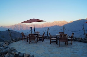 A terrace with stone flooring, wooden chairs, and tables, surrounded by a rope barrier, overlooking a mountain range. - Sama Wakan (Wakan)