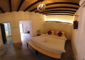 A bedroom with a wooden bench, a bed with pillows, a hanging light fixture, and a doorway leading to another room.
