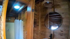 Une salle de bain rustique, avec des murs en bois, comprenant des toilettes blanches, un lavabo avec un miroir au-dessus et un espace douche.