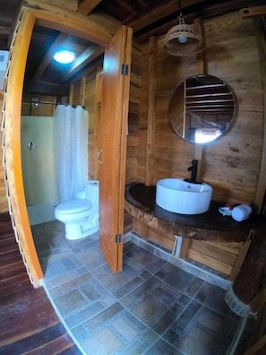 Baño de cabaña de madera con inodoro blanco, lavabo con espejo arriba y zona de ducha.