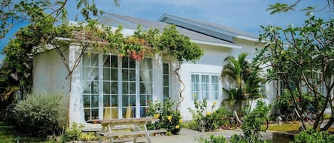 Una casa con un amplio ventanal, un banco de madera y un jardín con plantas y una bicicleta.