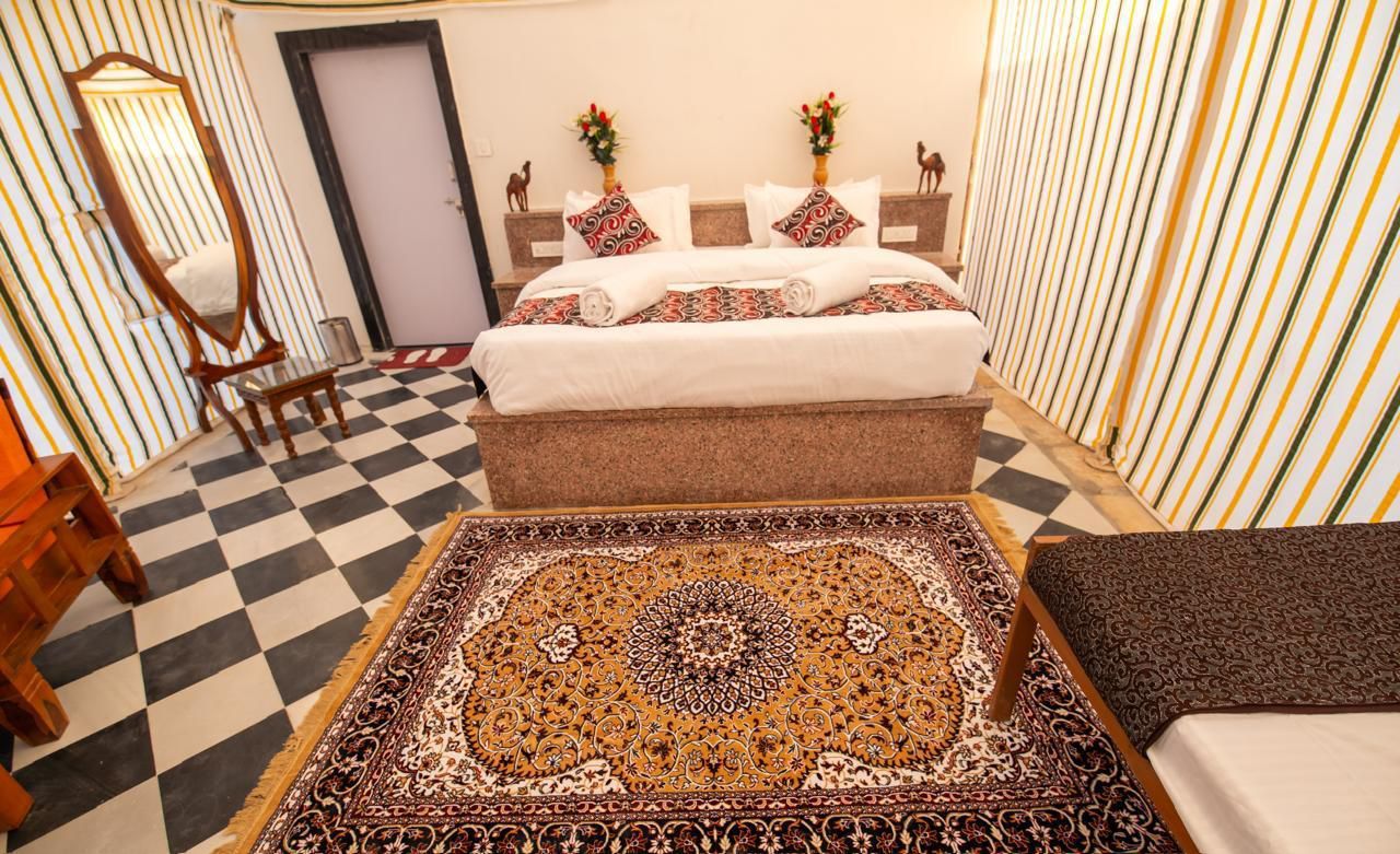 Una habitación de hotel con piso a cuadros, una cama grande con ropa de cama estampada, un área de descanso con un sofá, un espejo y elementos decorativos en las paredes.