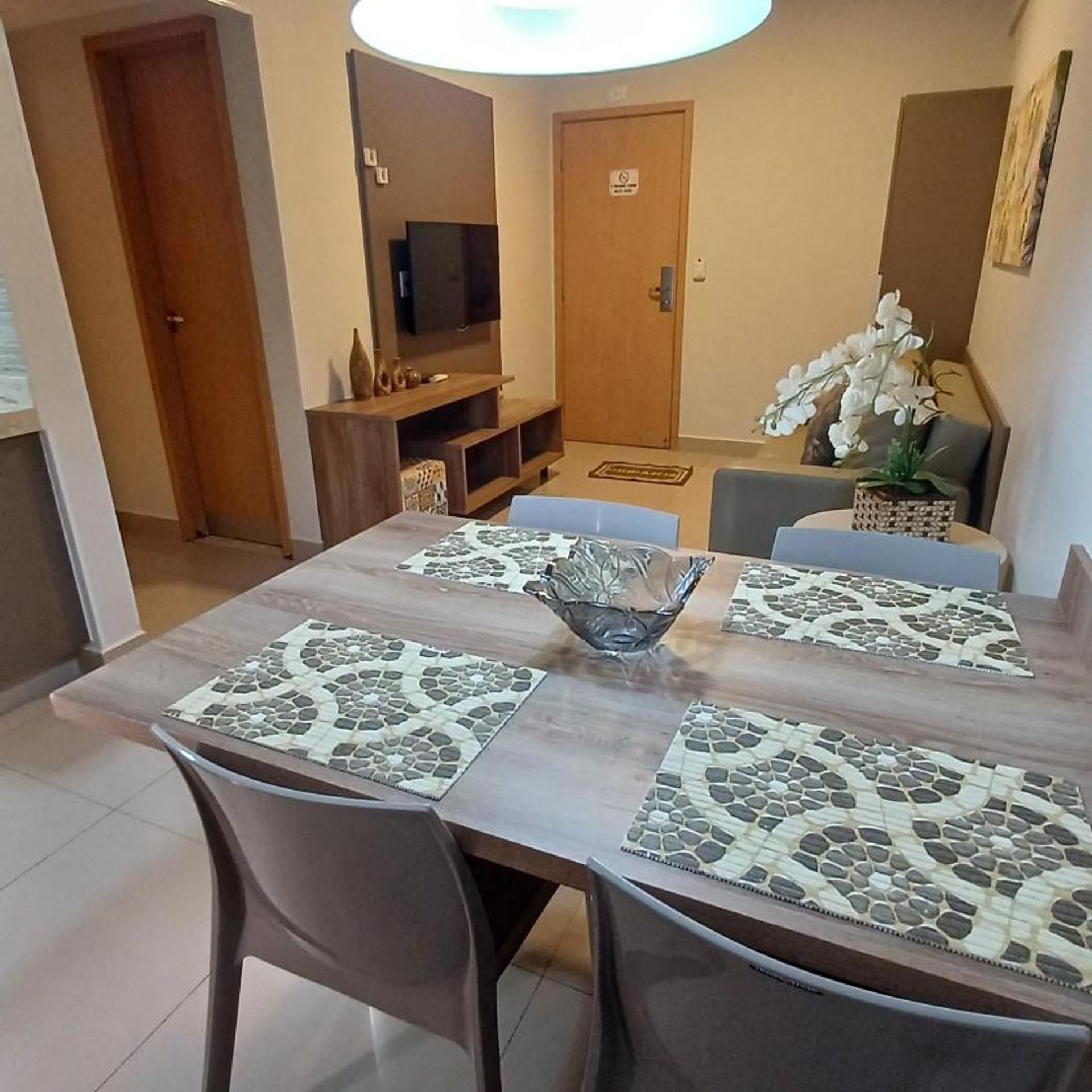 Uma sala de jantar com mesa de madeira, duas cadeiras, um centro de mesa em vidro e uma televisão ao fundo.