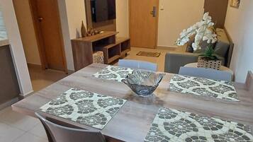 Uma sala de jantar com mesa de madeira, duas cadeiras, um centro de mesa em vidro e uma televisão ao fundo.
