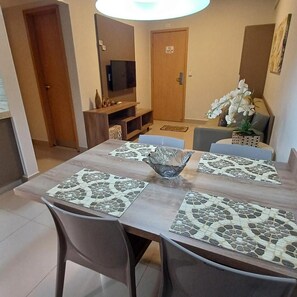 Un coin repas avec une table en bois, deux chaises, un grand saladier en verre au centre et un téléviseur en arrière-plan.