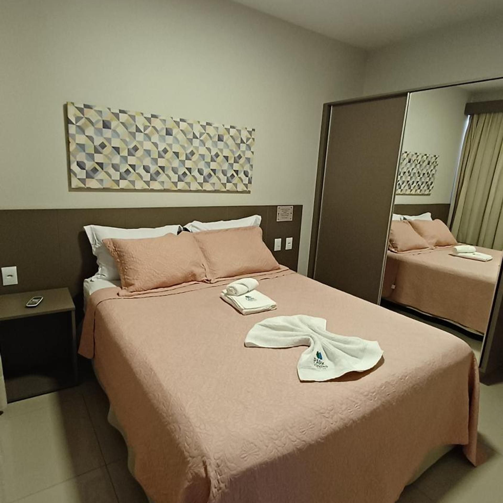 Quarto de hotel com cama, mesa de cabeceira, espelho e um ornamento geométrico na parede.