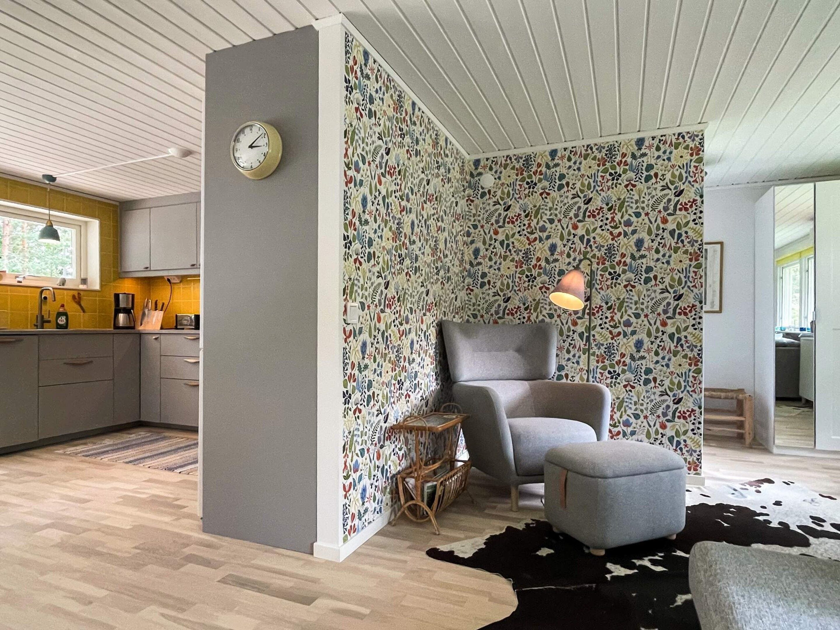 A modern living room with a patterned wallpaper, a grey armchair, a small ottoman, a clock, and a cowhide rug.