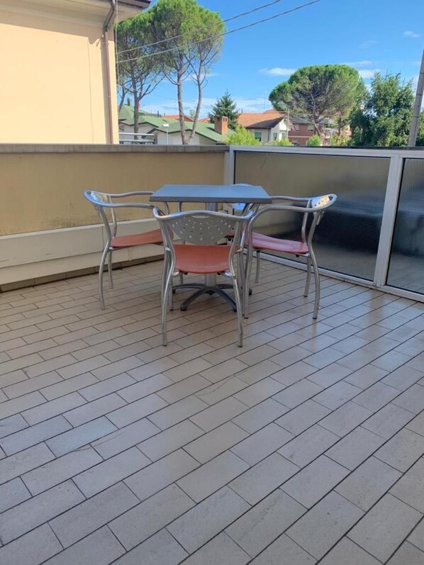 Pátio externo com mesa e cadeiras, piso de azulejos e vista para casas e árvores ao fundo.