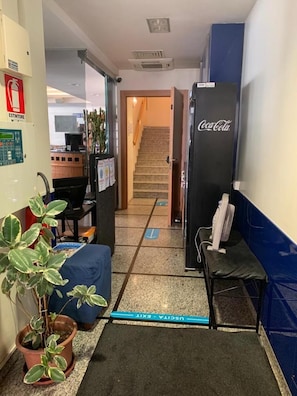 Corredor de hotel com escada, geladeira Coca-Cola, mesa com computador e planta em vaso.
