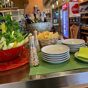 Disposição de buffet com diversas opções de comida, incluindo uma saladeira, garrafas de bebidas e pilhas de pratos.