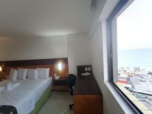 A hotel room with a bed, a desk, and a window with a view of the city and the sea. - Natal Plaza Requinte Conforto E Vista Para O Mar (Natal)