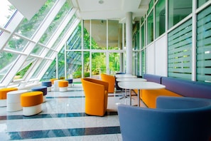 Ein modernes Interieur mit einem Schachbrettmuster-Boden, Sitzbereichen mit orange- und blauer Möbel und großen Fenstern mit Blick auf Grünflächen.