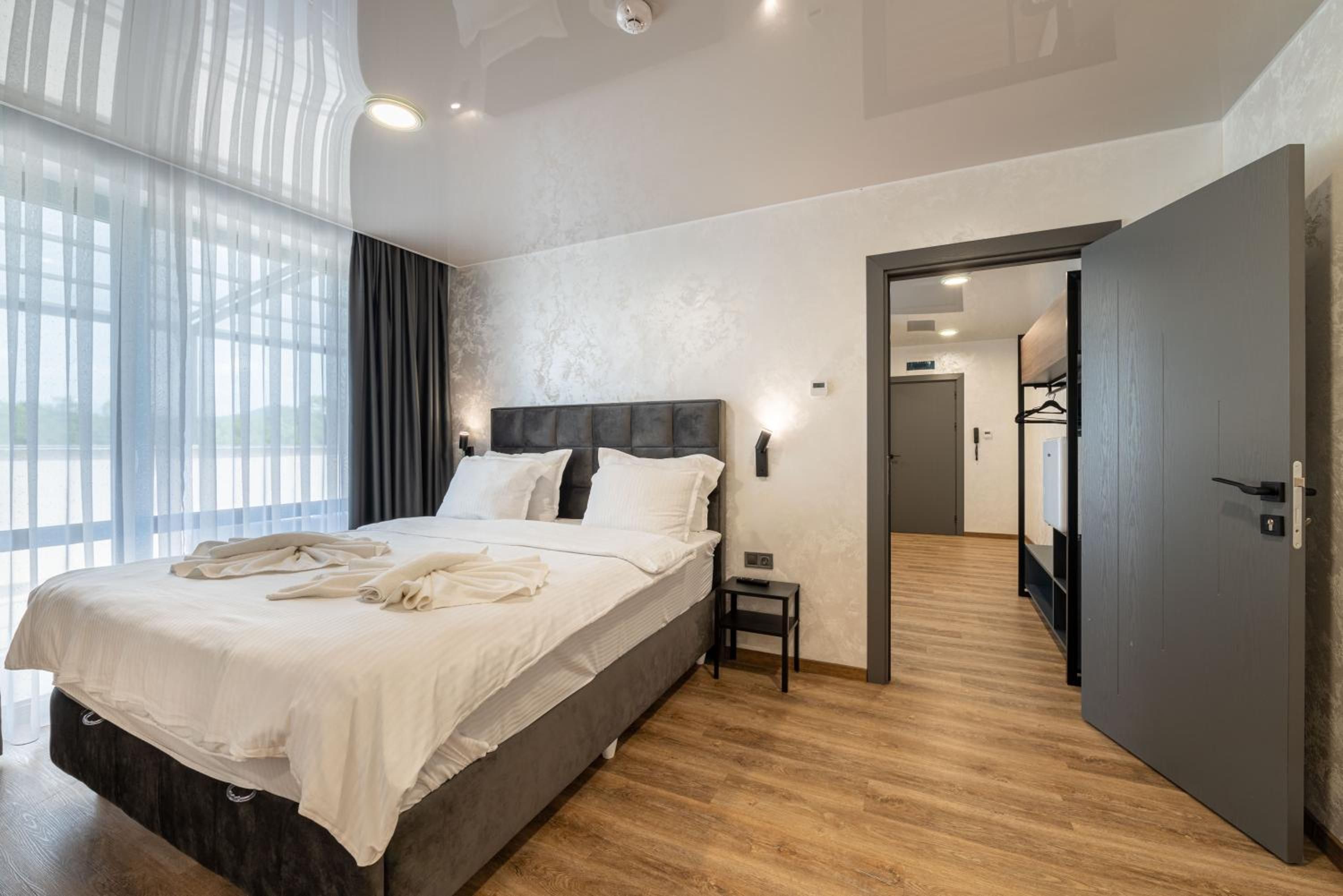 A modern hotel room with a large bed, a bedside table, a wardrobe, and a view through a window with sheer curtains.