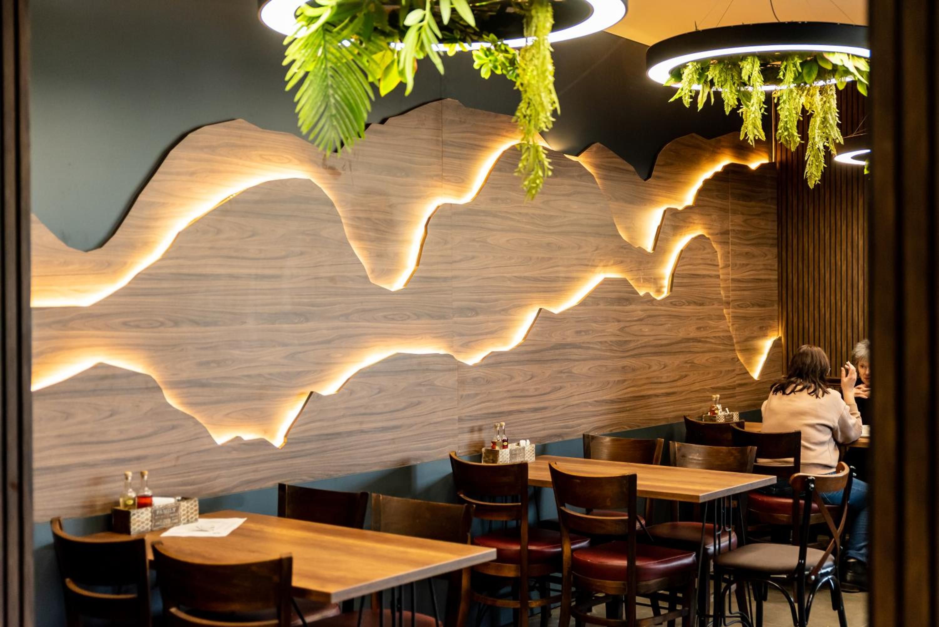 A restaurant interior with wooden tables and chairs, a wall with a wave-like light design, and hanging plants.