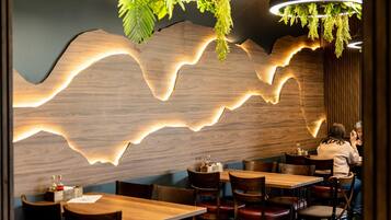 Un interior de restaurante con mesas y sillas de madera, una pared con un diseño de luz en forma de ola, y plantas colgantes.