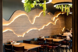 A restaurant interior with wooden tables and chairs, a wave-like light feature on the wall, and hanging greenery. - Hotel Royal Bath (Karlovo)