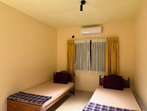 A room with two single beds, a window with yellow curtains, an air conditioning unit, and a ceiling light. - Don Cyril's Nature Bungalow (Galle)