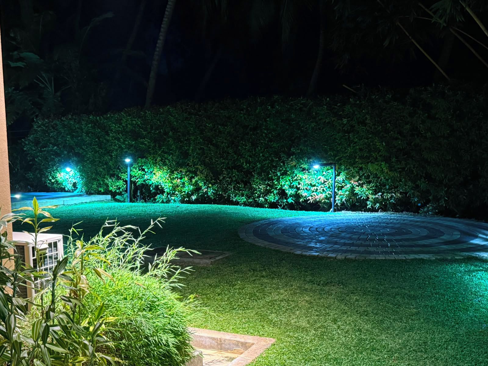 A well-lit garden area with a circular paved path, surrounded by neatly trimmed hedges and illuminated by street lamps.