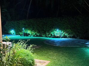 A well-lit garden area with a circular paved path, surrounded by neatly trimmed hedges and illuminated by street lamps.