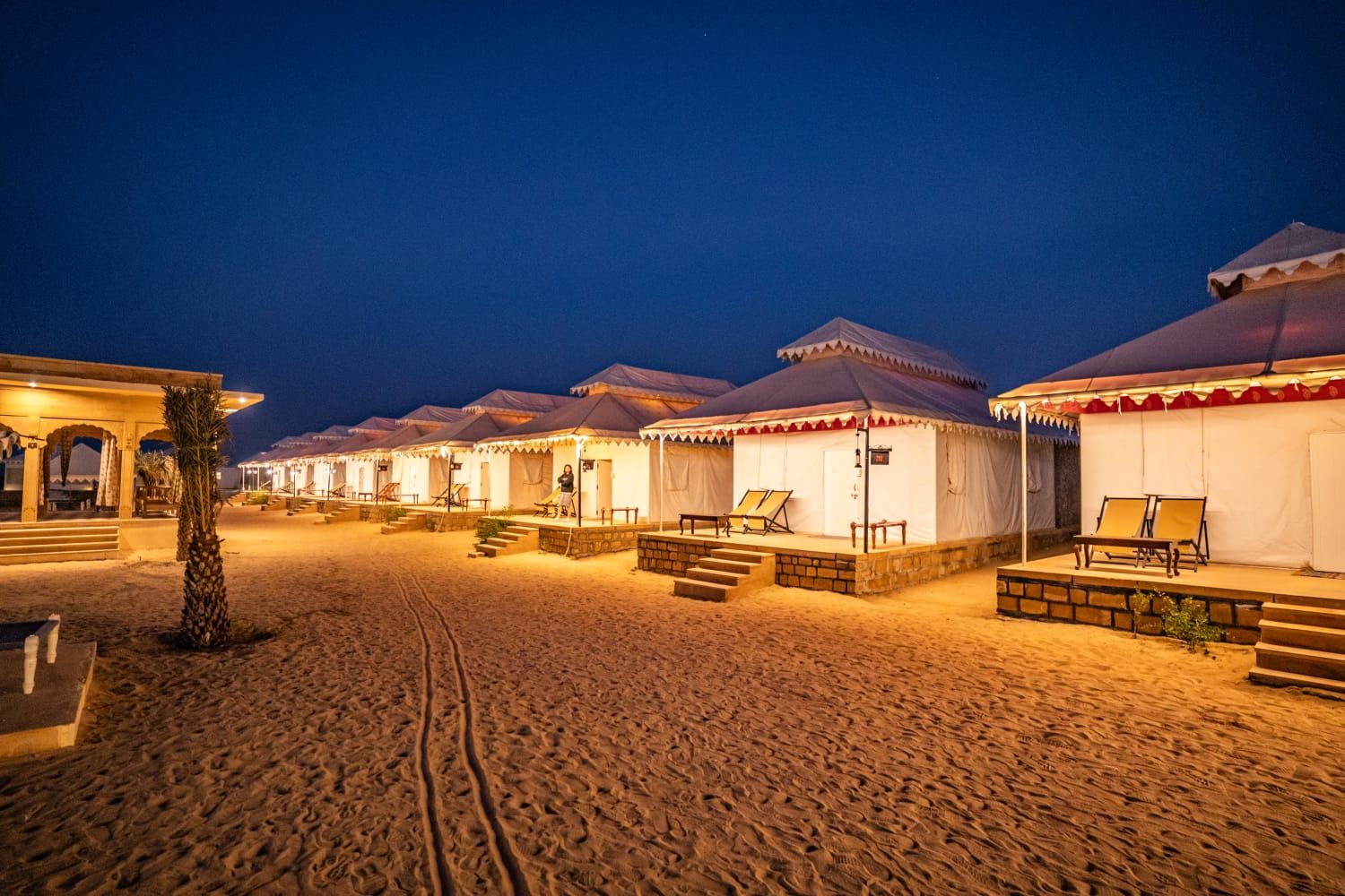 A row of tents with steps leading to the entrance, a palm tree, and a sandy ground.