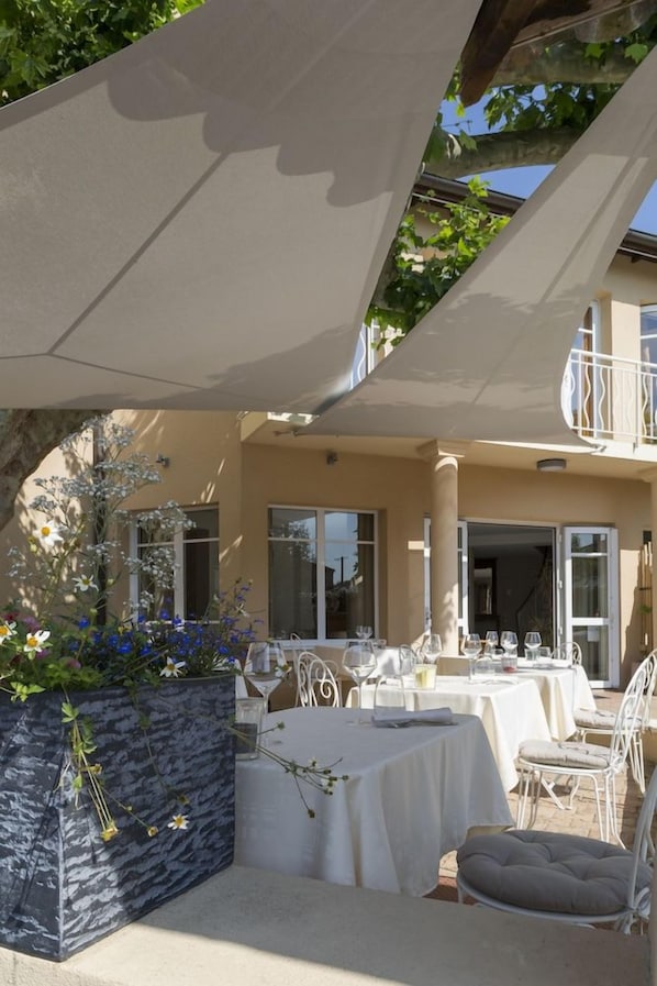 An outdoor dining area with tables and chairs, a planter with flowers, and a building in the background. - Auberge De Clochemerle (Vaux-en-Beaujolais)