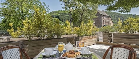 Un espace de restauration en plein air avec une table dressée pour deux personnes, comprenant une assiette de croissants, une théière, des tasses et un verre de jus d’orange.