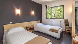 A hotel room with two beds, a nightstand with a lamp, a chair, a window with a view of trees, and a radiator. - Hotel Eco - A75 (Banassac)