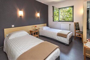 Habitación de hotel con dos camas, una mesita de noche con lámpara, una silla, un ventanal con vistas a árboles y un radiador.
