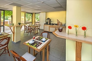 Un área de comedor con mesas y sillas, una barra con caja registradora, y una pared decorada con arreglos florales.