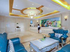 Un vestíbulo de hotel con una recepción, una araña, un cuadro grande y mobiliario tapizado en azul.