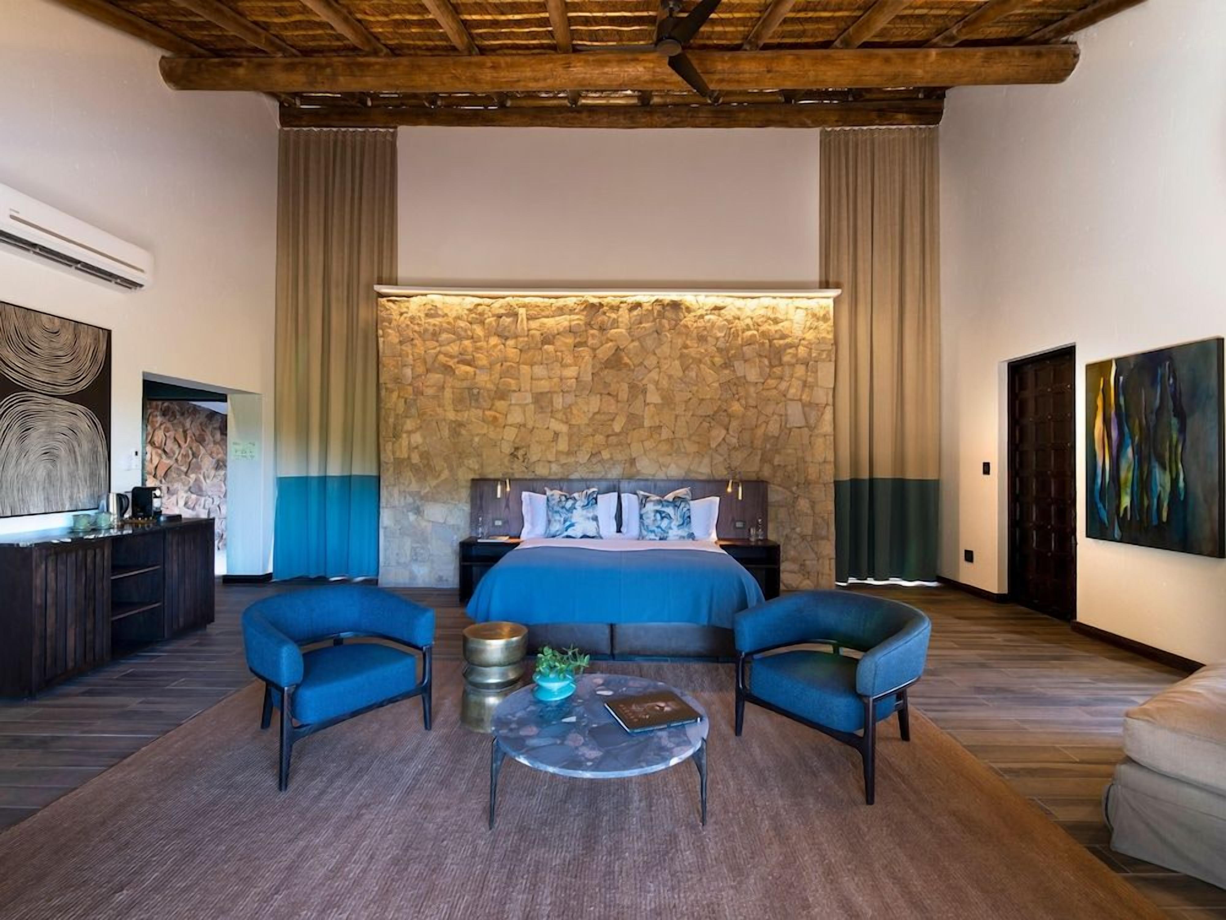 A bedroom with a stone wall, a bed with blue bedding, two blue armchairs, a coffee table, and a TV on the left.