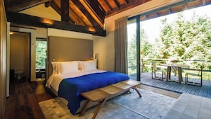 Quarto com cama grande, um banco de madeira, varanda com mesa e cadeiras, e vista para árvores.
