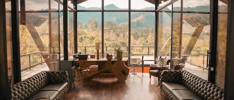 A spacious room with a large window offering a view of a mountainous landscape. There is a wooden table with chairs and a sofa.