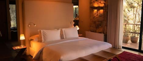 A bedroom with a large bed, bedside lamps, a wooden bedside table, a red rug, and a stone wall.