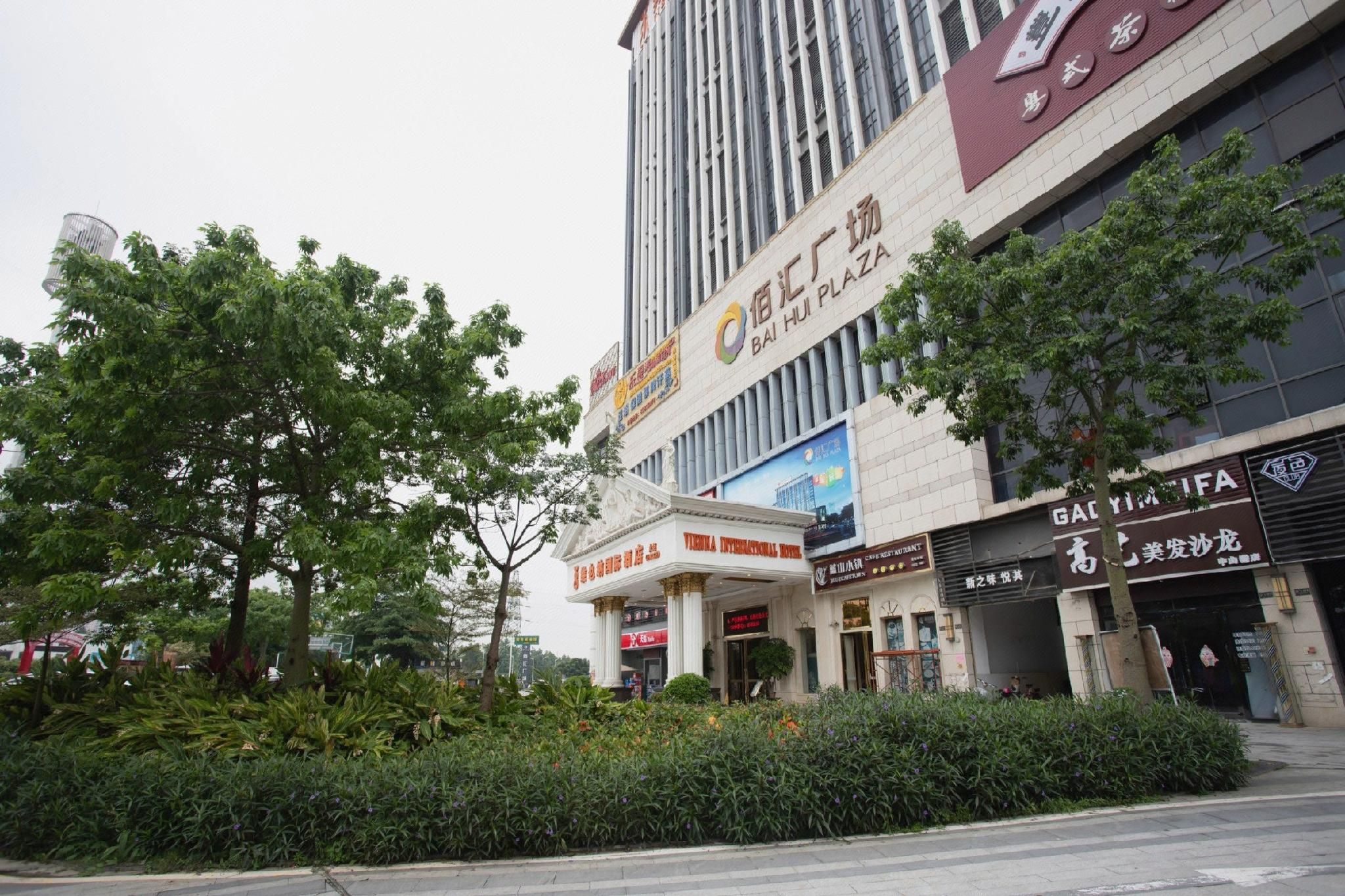 Un hotel con un letrero llamativo, un gran árbol en primer plano y varios comercios.