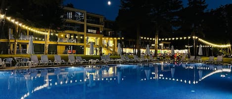 A swimming pool with lounge chairs and umbrellas, surrounded by a building with multiple floors and string lights.