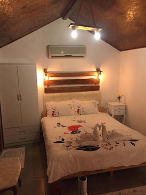 Quarto com teto de madeira, cama com roupa de cama florida, mesa de cabeceira com abajur e guarda-roupa.
