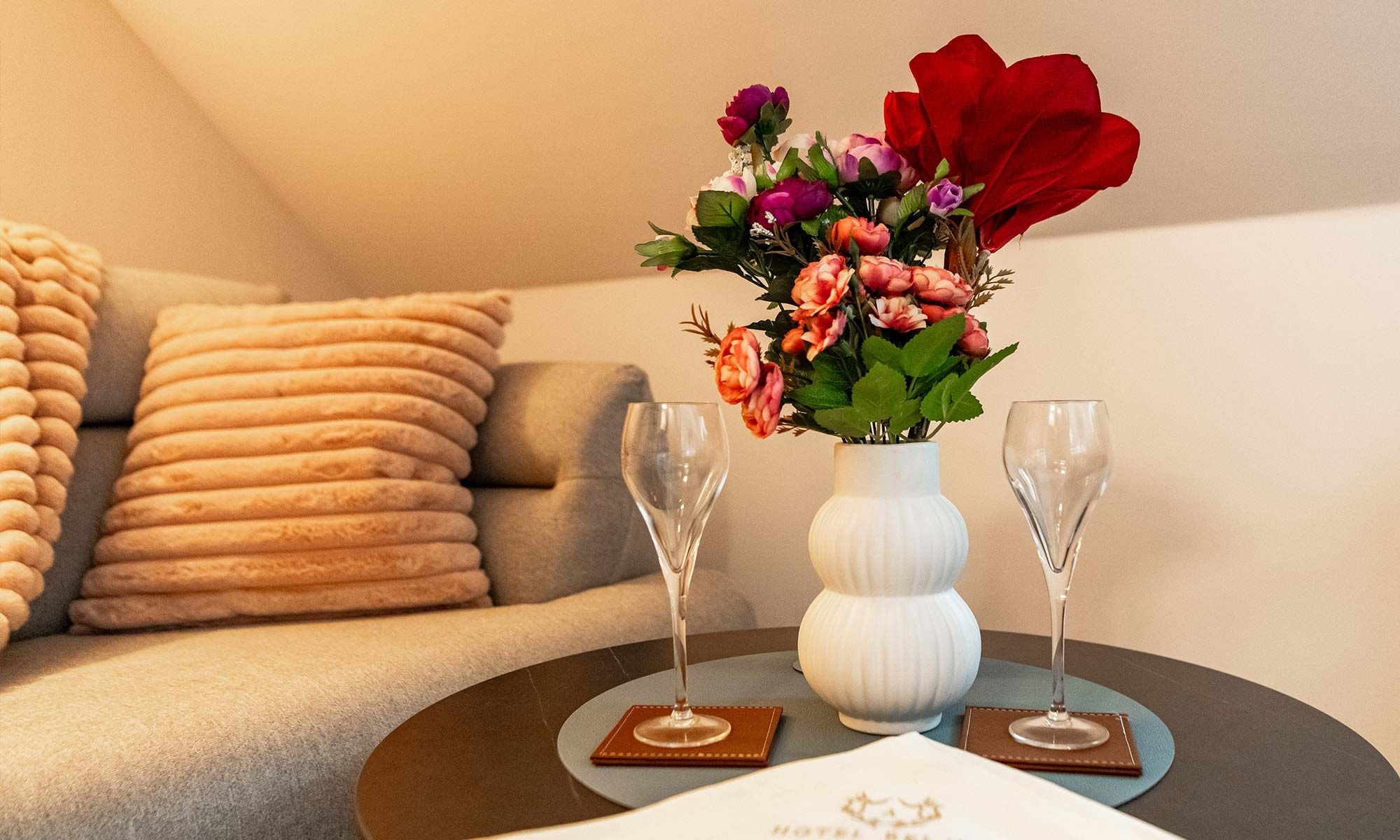 Una habitación con un sofá, un jarrón blanco con flores y dos vasos sobre una mesa redonda.