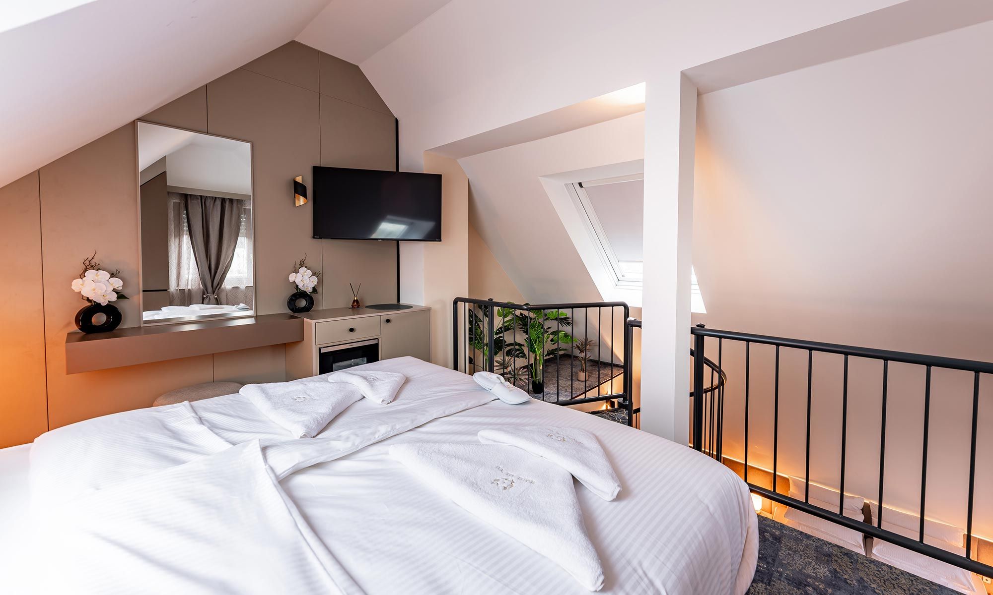 A modern hotel room with a large bed, a flat-screen TV, a small kitchenette, and a balcony with plants.