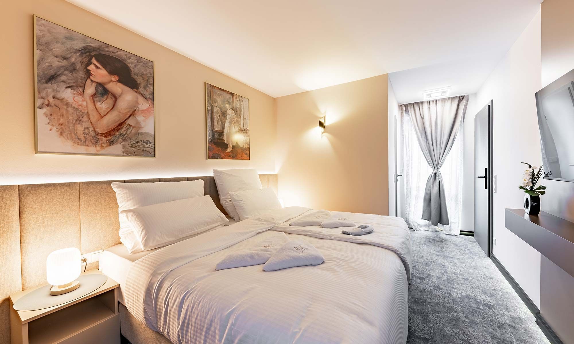 A hotel room with a large bed, a bedside table with a lamp, a painting on the wall, and a window with curtains.
