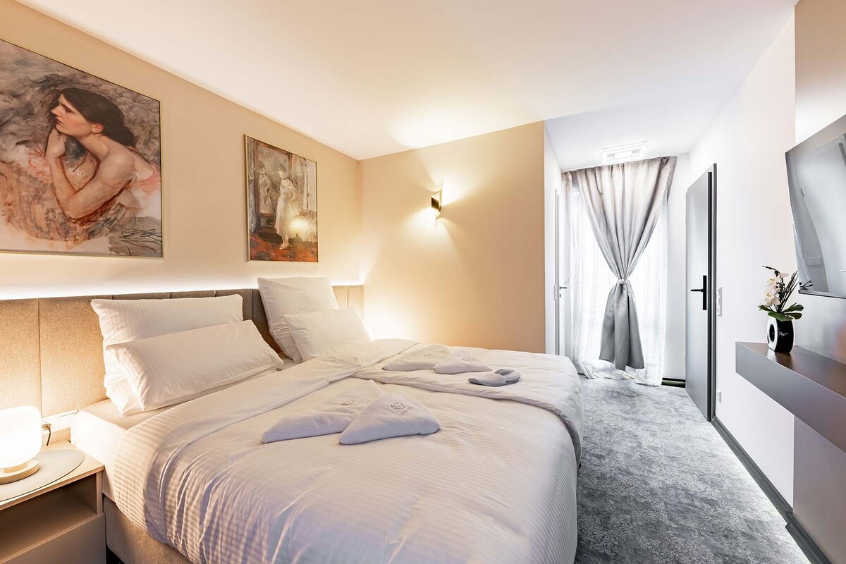 A hotel room with a large bed, a bedside table with a lamp, a painting on the wall, and a window with curtains.
